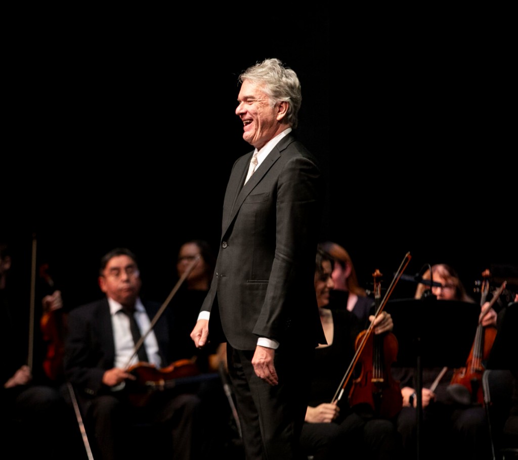 Dr. Brian Stone smiling on stage with orchestra musicians behind him